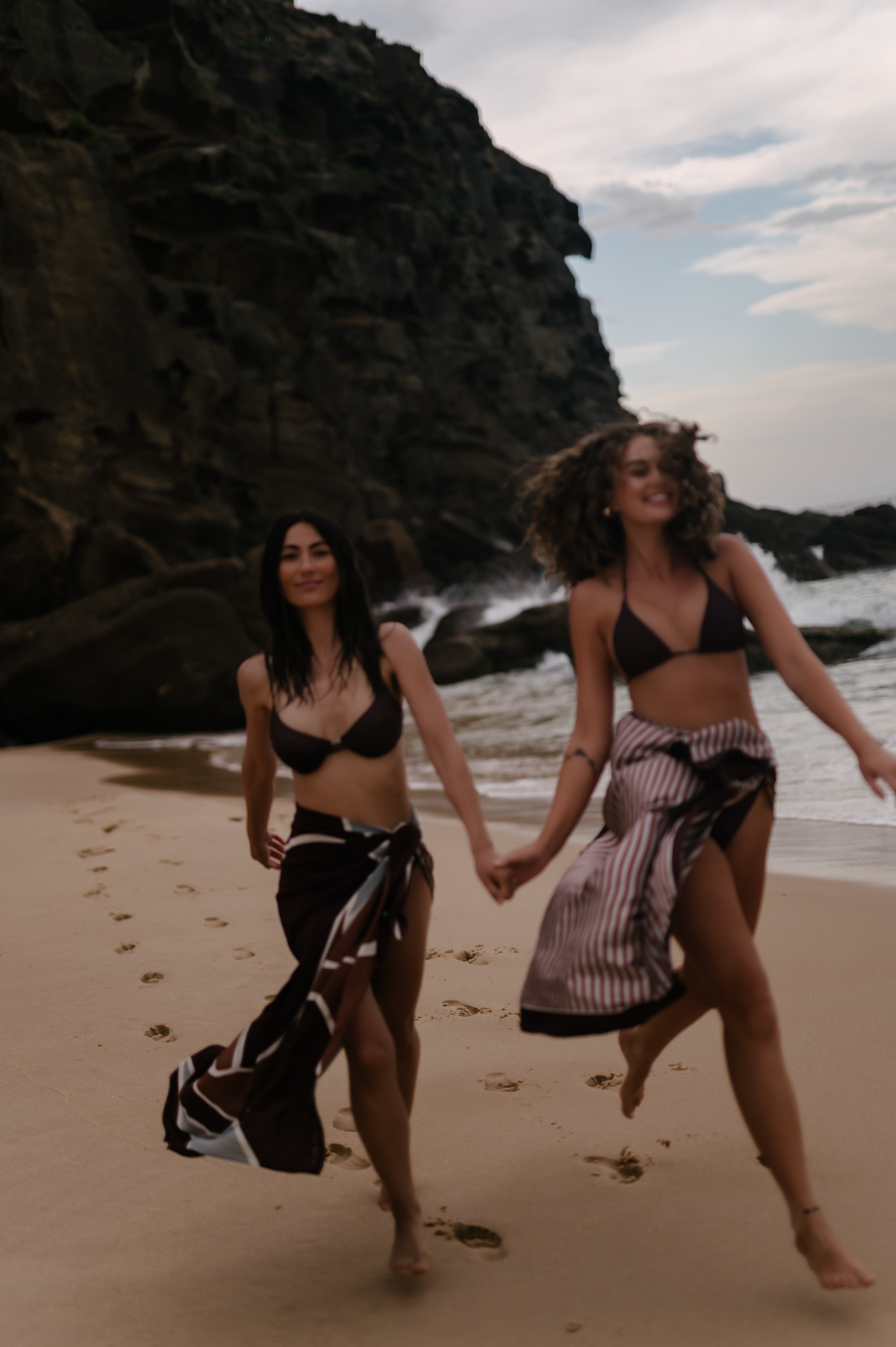 Two women walking on a beach holding hands with rocky cliffs and ocean in the background. They are wearing Novo Pareo sarong wraps made from 100% silk.