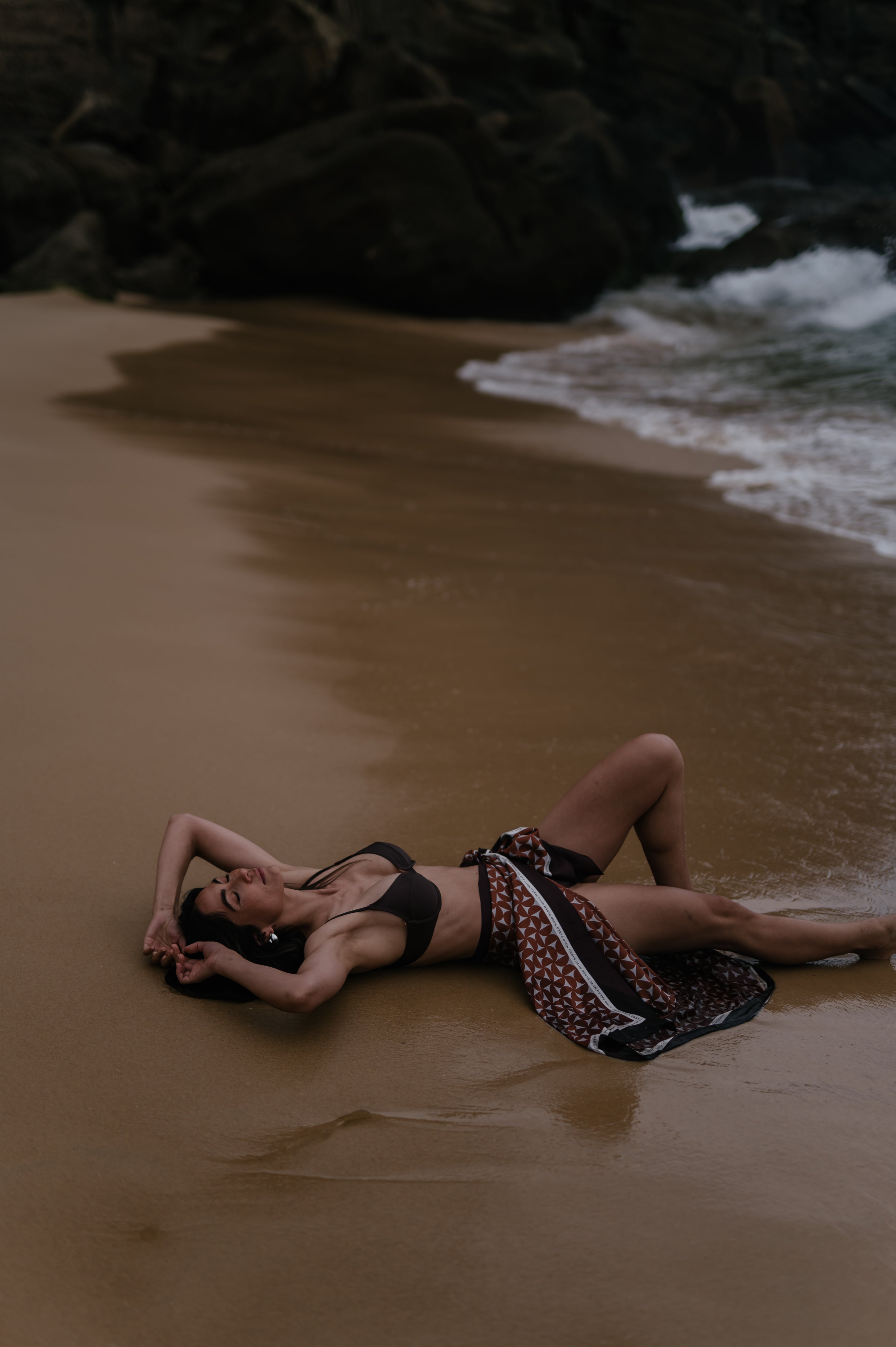 Model lying on a beach with waves in the background. She is wearing a Novo Pareo Silk Wrap Sarong. Australian Resort Wear.