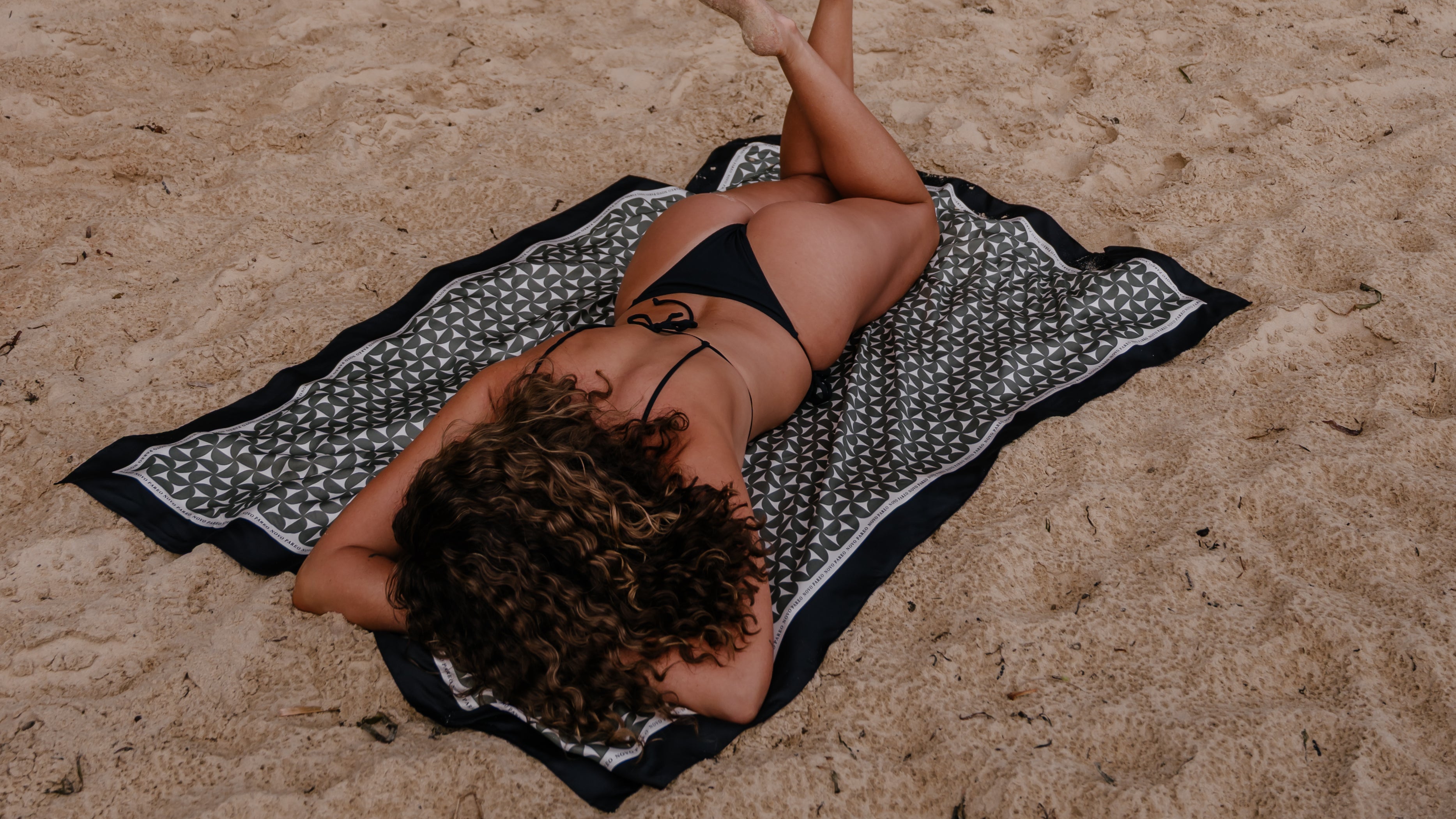 Woman lying on the sand using a black geometric Novo Pareo sarong as a beach mat, with natural rock formations in the background.