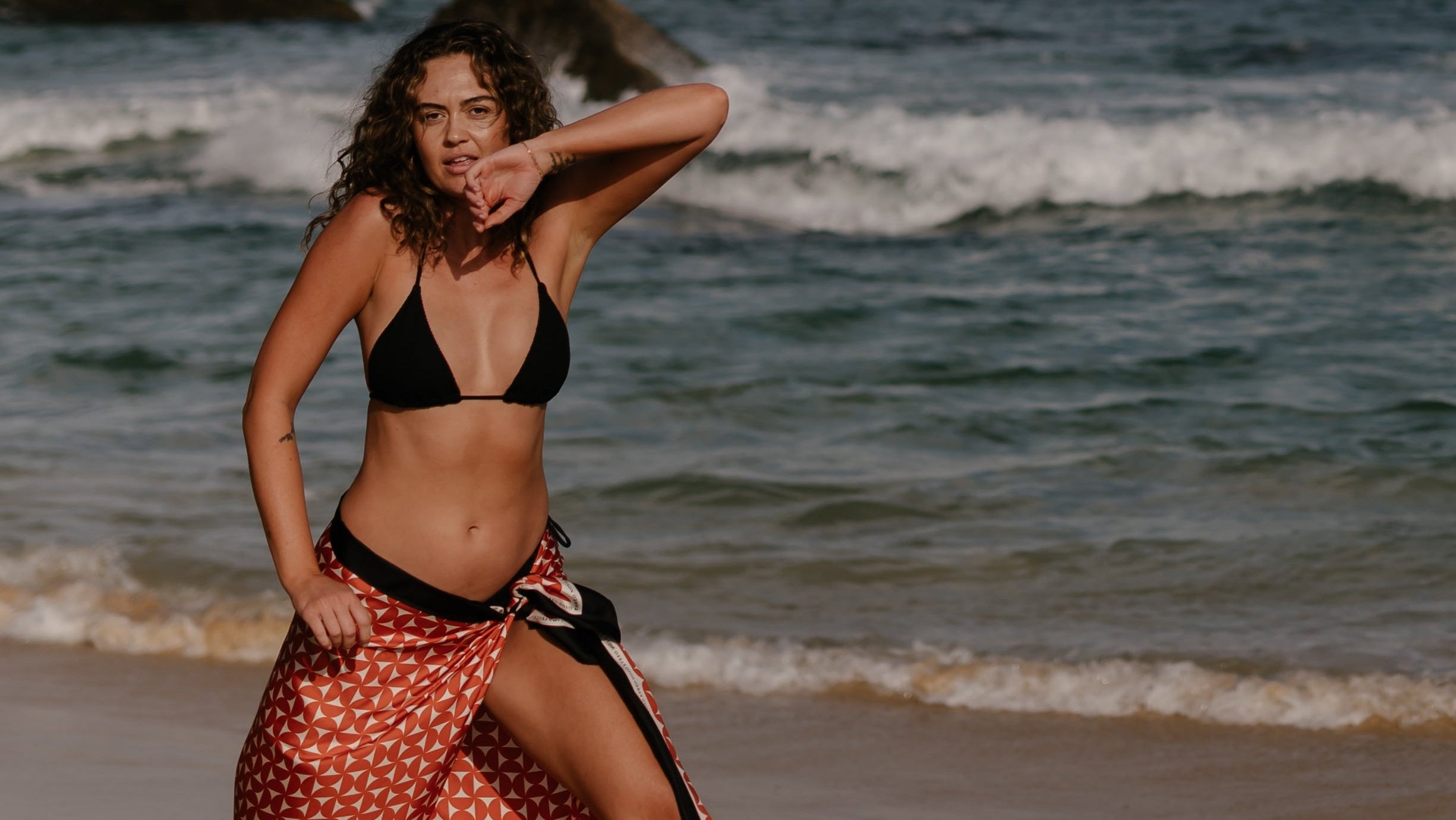 A woman wearing the Blaire terracotta 100% silk pareo wrap sarong by Novo Pareo standing on the beach, with waves and rocky coastline in the background. Australian luxury resort wear.