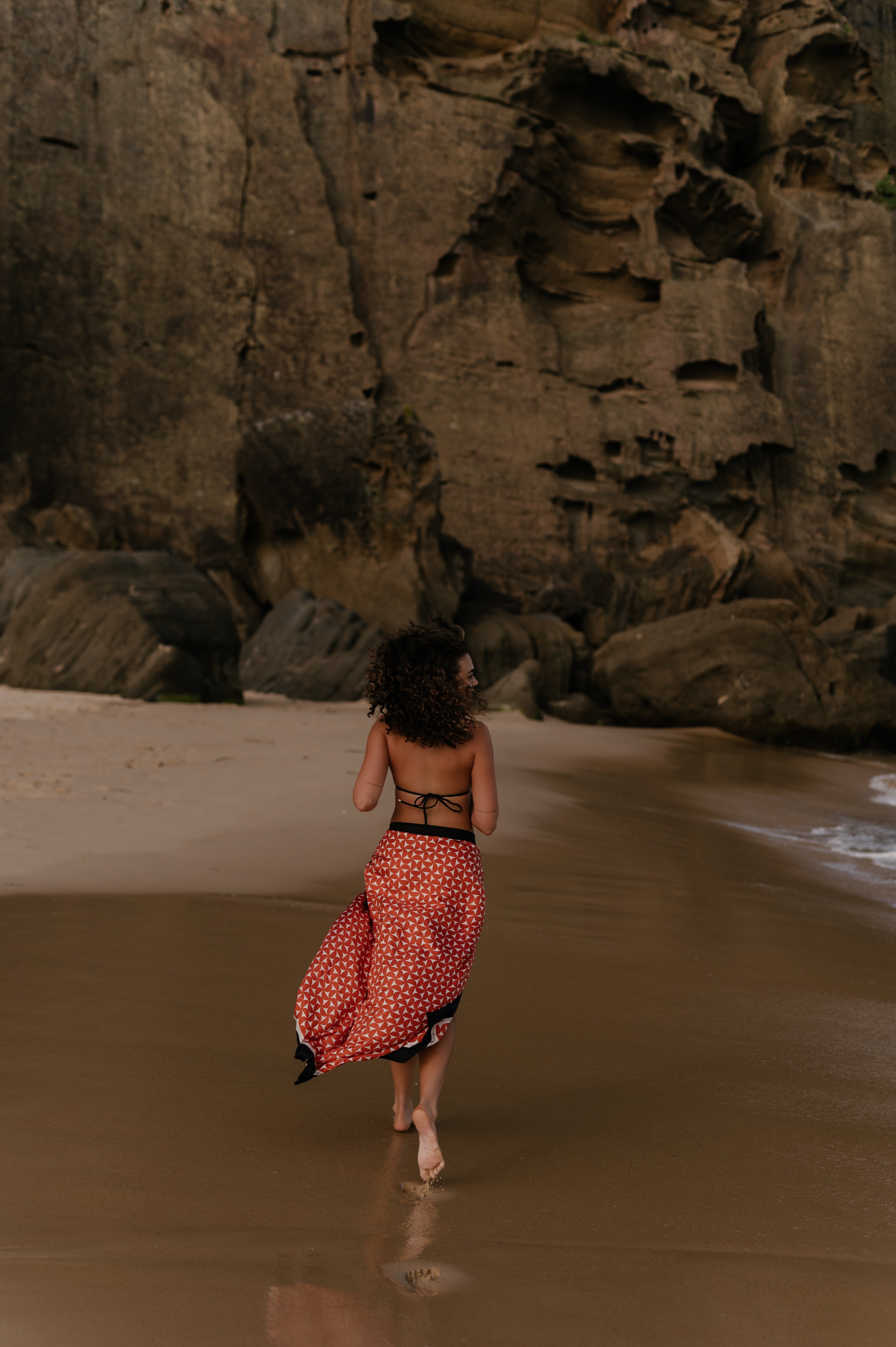 Woman walking along a sandy Australian beach wearing a red geometric Novo Pareo sarong, with rocky cliffs in the background.