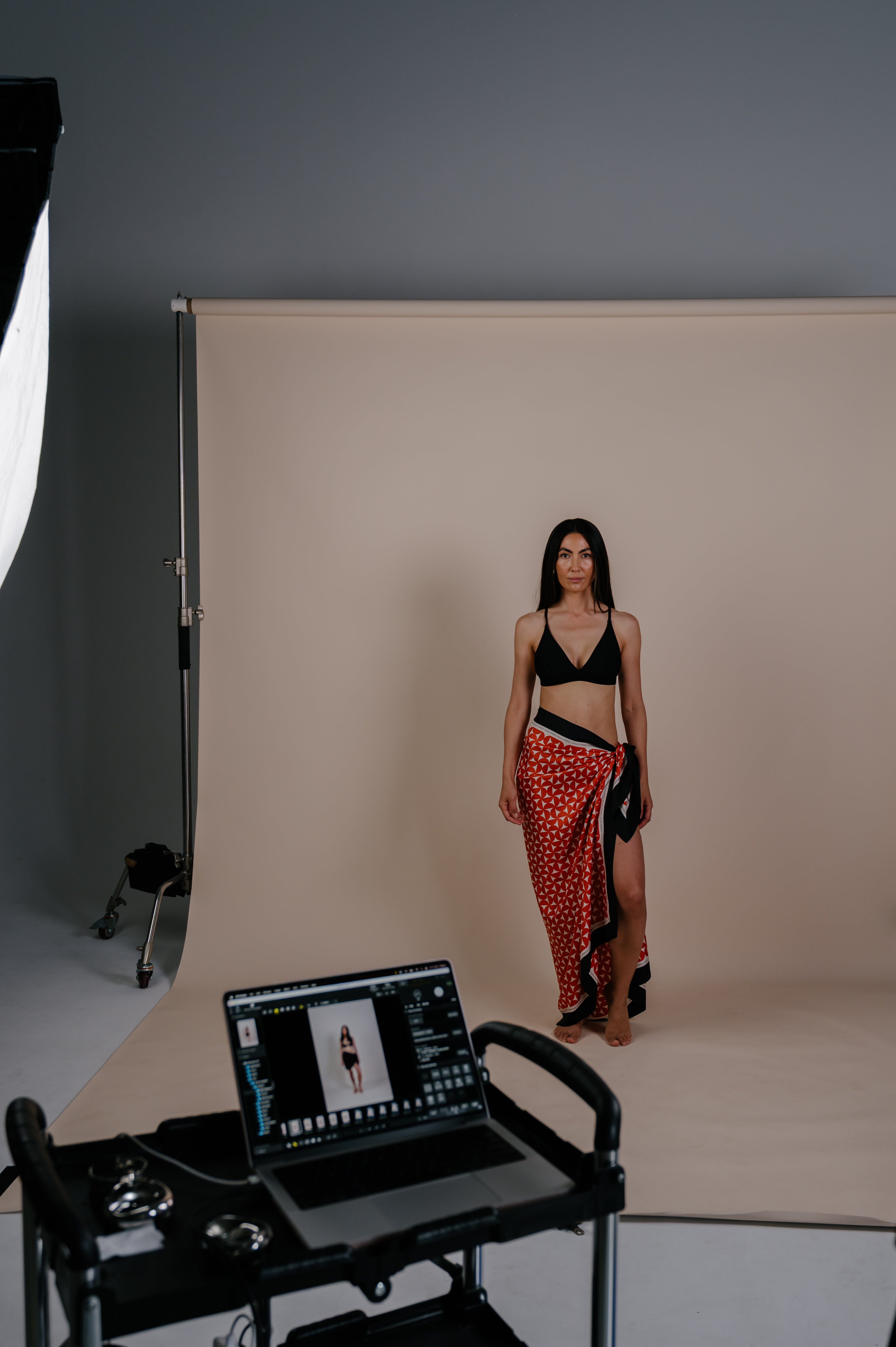 Model standing in a studio wearing a red geometric Novo Pareo sarong wrap and black swim top, with a neutral backdrop and photoshoot equipment visible.