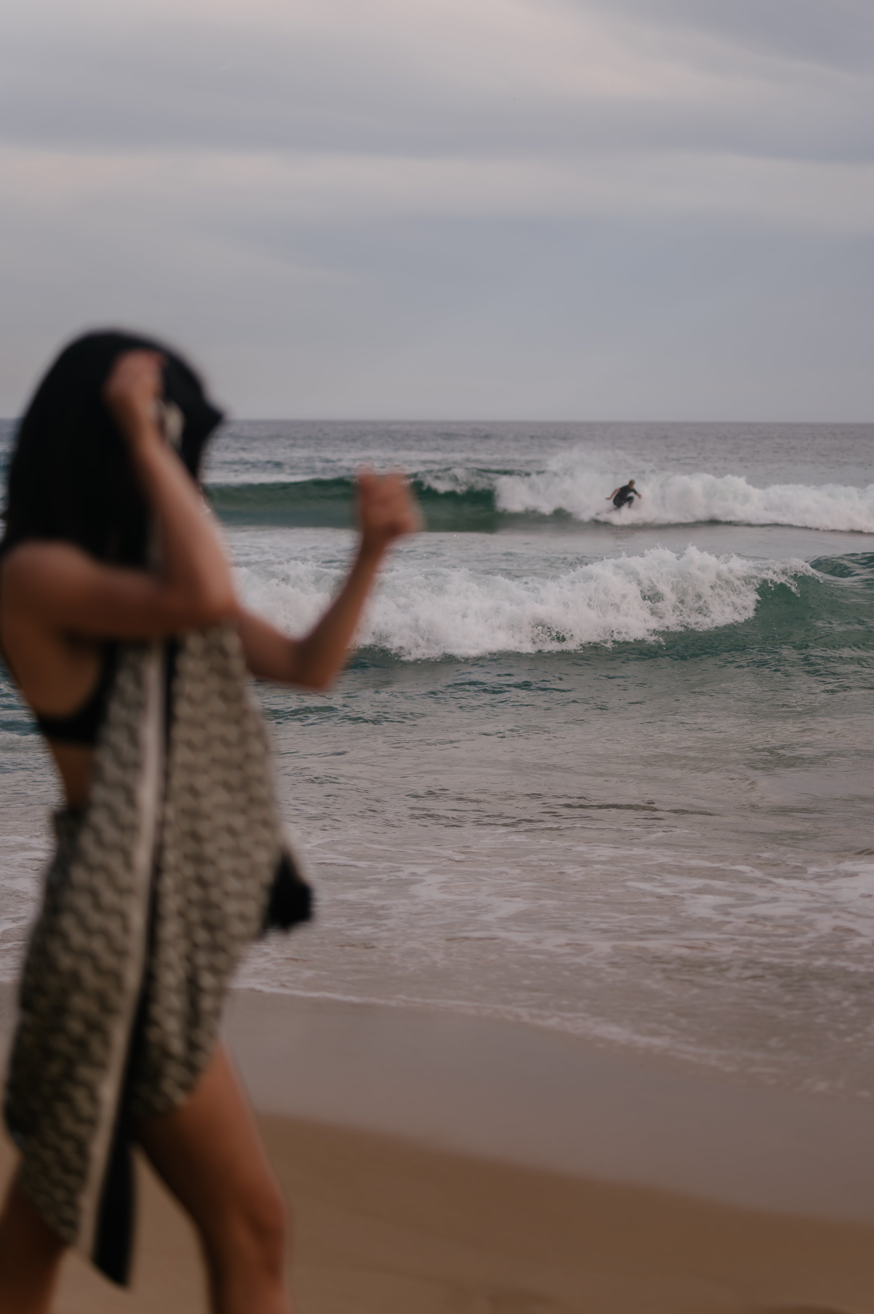 Person on a beach with ocean waves and another person surfing in the distance. She is wearing a Novo Pareo Sarong Wrap made of 100% silk.
