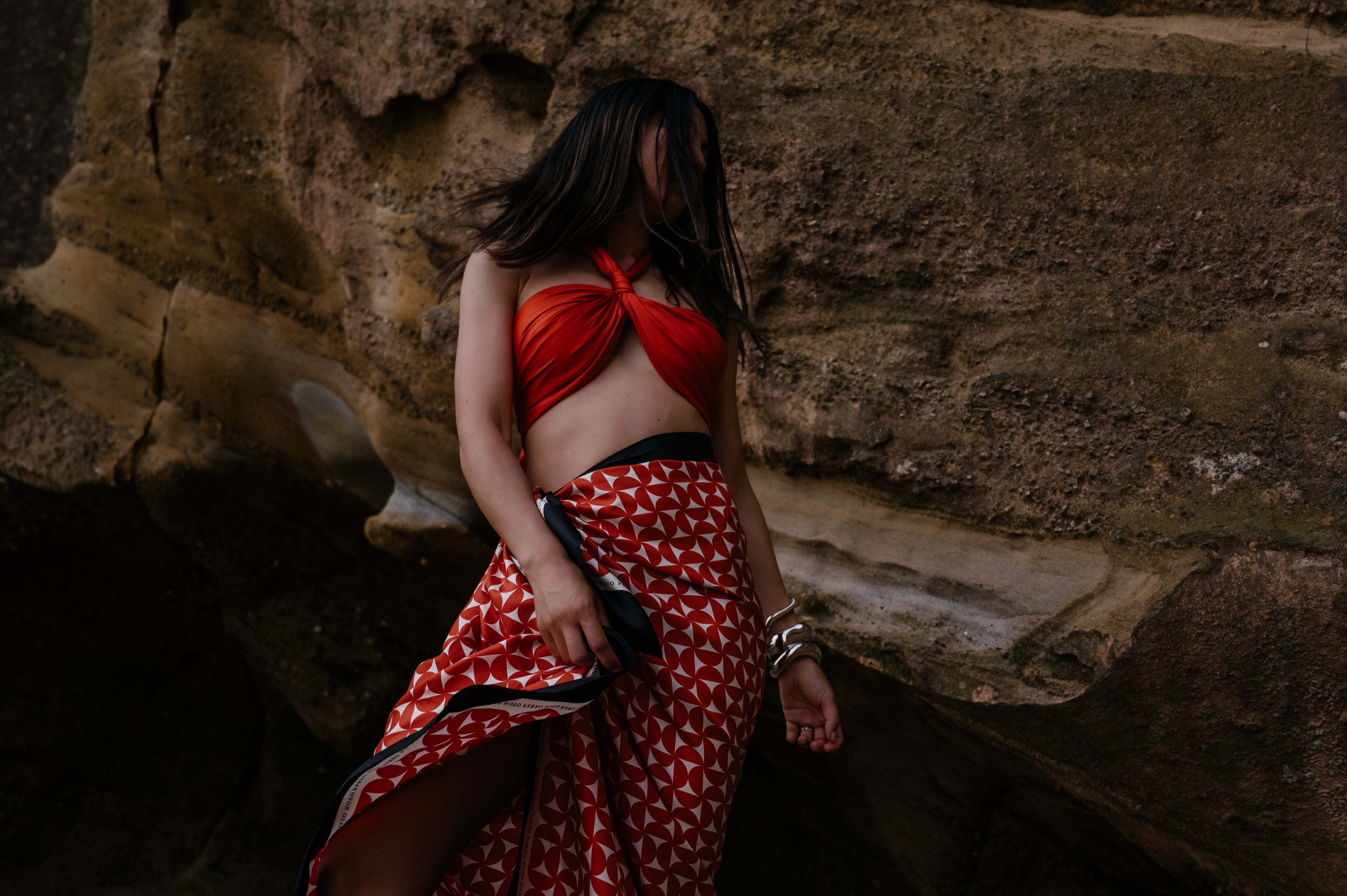 Woman in a red pareo sarong wrap and patterned skirt standing against a stone wall.