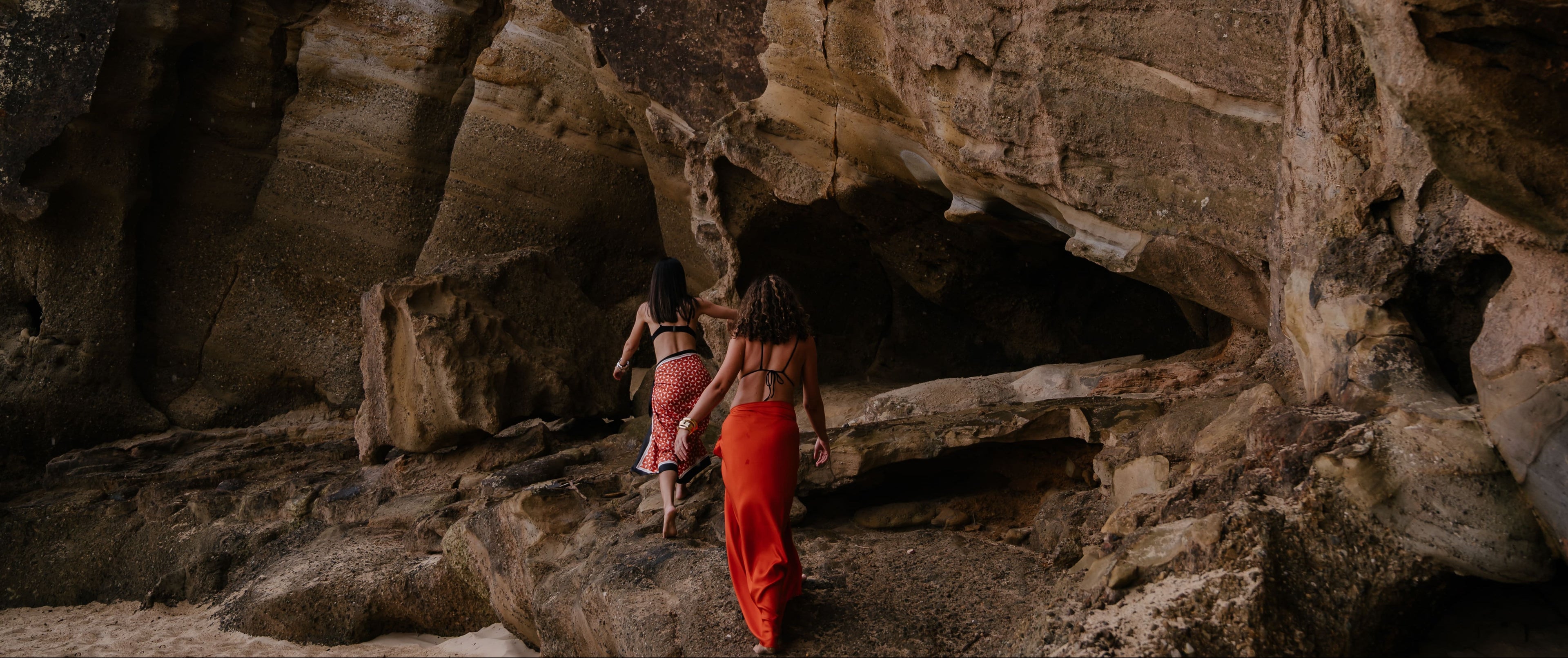 Two women walking along large coastal rock formations, wearing Novo Pareo Sarong wraps in red tones during a beachside lifestyle shoot.