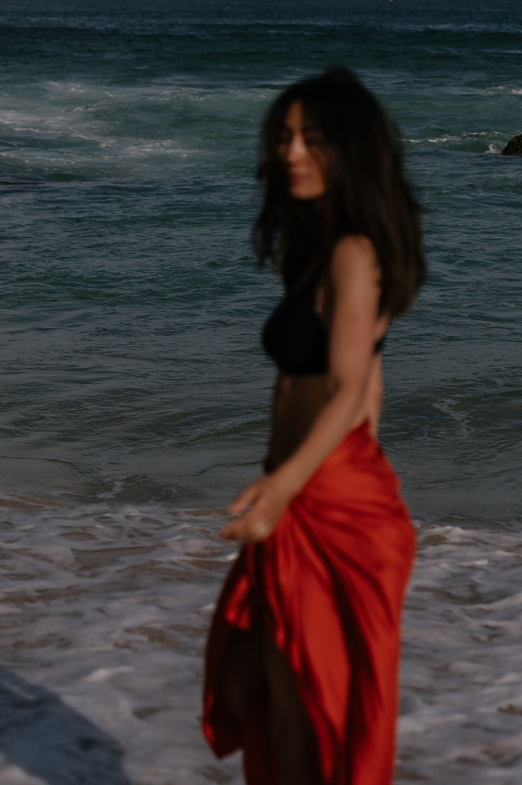 Woman in a red sarong standing on a beach with ocean waves in the background.