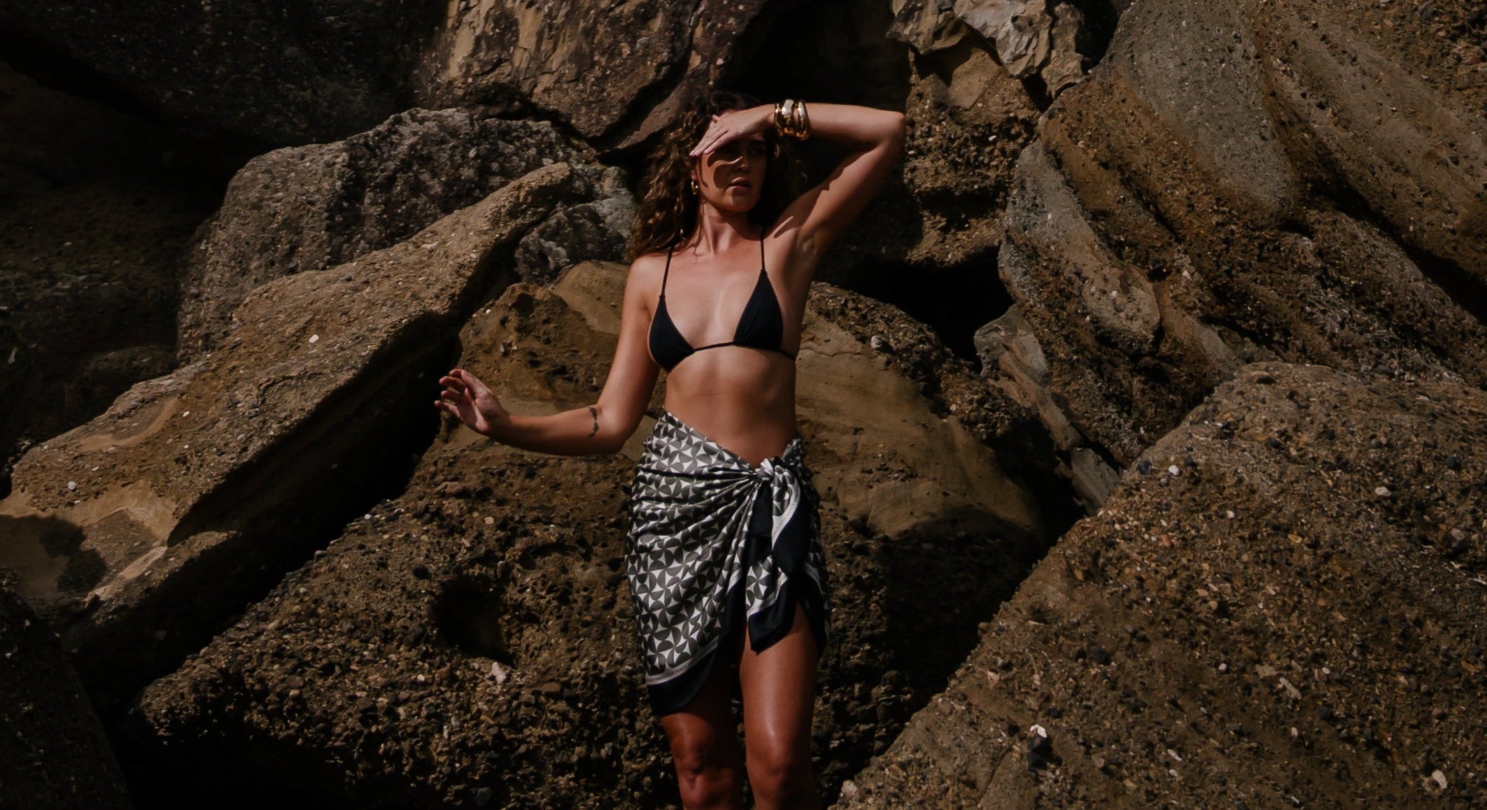 Model wearing the Solenne 100% silk pareo sarong wrap by Novo Pareo, standing against textured Australian coastal rocks. Luxury resort wear photography showcasing elegant, minimalist beach style.