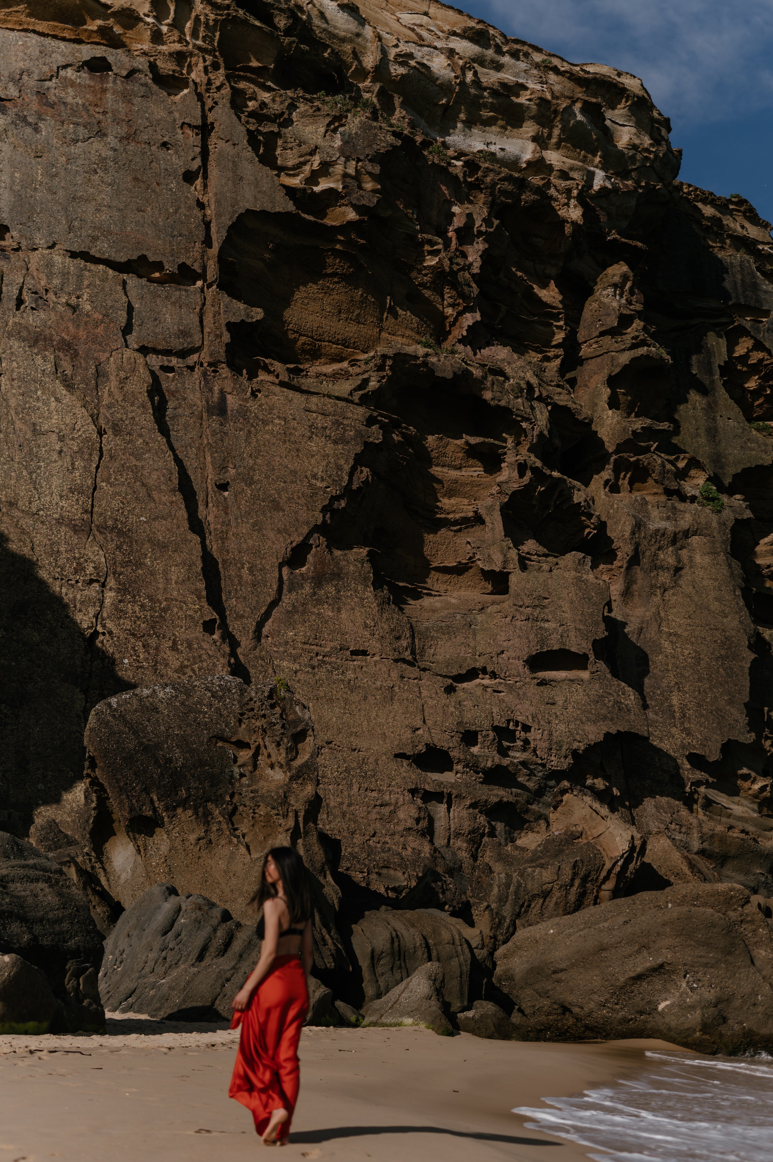 Person in a red dress standing on a beach with large rocks and ocean waves in the background. Novo Pareo silk sarong, wrap, resortwear.