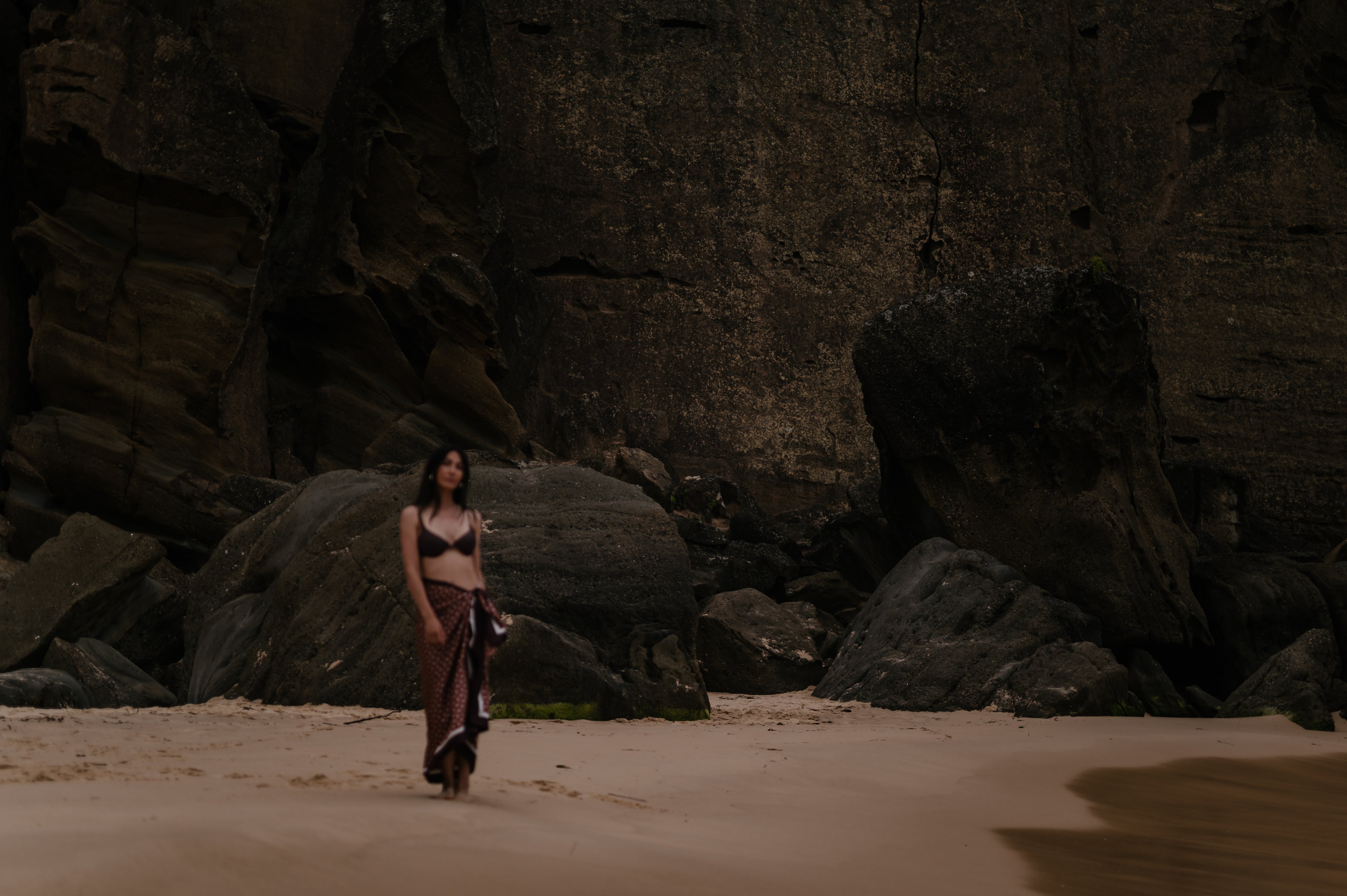 Model standing on a sandy beach with large rocks in the background. She is wearing a chocolate brown and dusty light blue Novo Pareo Sarong from the Intent Collection. 